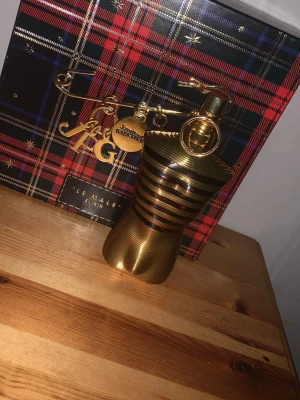 Jean Paul Gaultier Le Male Elixir 90ml kvar  - Lyxig parfymflaska i guld från Jean Paul Gaultier, Le Male Elixir. Flaskan har en ikonisk kroppsliknande form med horisontella ränder och metallisk finish. Kommer i en snygg presentförpackning med tartanmönster och gulddetaljer.
