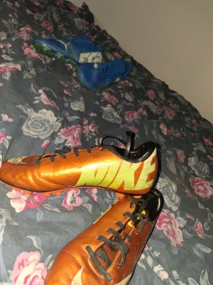 Nike orangea fotbollsskor - Säljer ett par orangea fotbollsskor från Nike med stora gula loggor på sidorna och svarta detaljer. Skorna har snörning och dobbar i orange och svart för bra grepp på planen. Perfekta för dig som vill sticka ut på matchen!