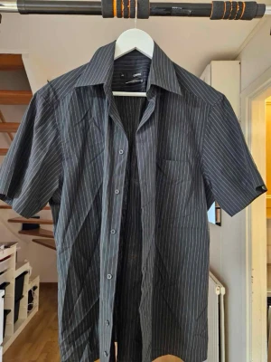 Svart kortärmad skjorta från Conwell - Snygg svart skjorta från Conwell med tunna vita ränder. Skjortan har korta ärmar, klassisk krage och bröstficka. Tillverkad i 65% polyester och 35% bomull vilket gör den både lätt och bekväm. Perfekt för en stilren look.