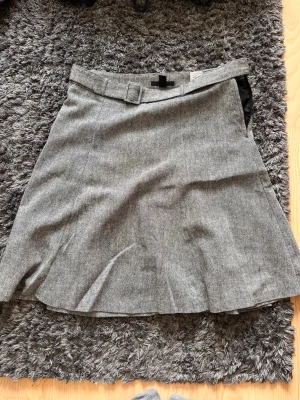 Grå A-linjeformad kjol med bälte - Snygg grå kjol i A-linjeform med tillhörande bälte i samma tyg. Kjolen har en klassisk look och är tillverkad i ett mjukt, vävt material som ger en stilren känsla. Perfekt att matcha med stickad tröja eller skjorta för en trendig outfit.