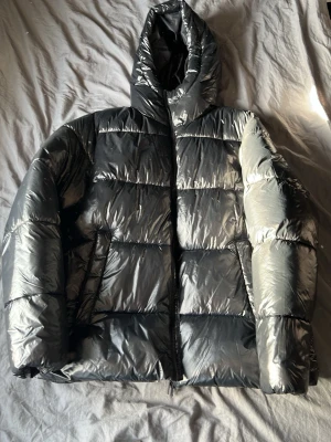 Silvrig pufferjacka från 157 - Säljer en riktigt fet silvrig pufferjacka från 157 med glansig finish och huva. Jackan har breda kanaler, dragkedja framtill och två sidofickor. Perfekt för kalla vinterdagar och ger en clean, modern look.