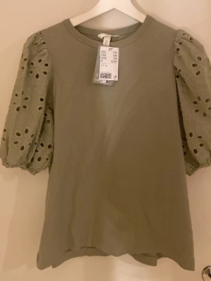 Grön topp med broderade puffärmar - Snygg ljusgrön topp från H&M med puffiga, broderade kortärmar i bomull. Ärmarna har hålmönster och ger en romantisk vibe. Rund halsringning och rak passform. Perfekt för dig som vill ha något extra till din outfit.