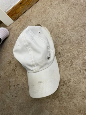 Vit keps med metallring - Säljer en stilren vit keps med böjd skärm och en liten metallring som detalj på sidan. Kepsen har klassisk sexpanelsdesign och justerbar rem bak. Perfekt accessoar för en clean och avslappnad look.