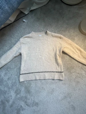 Beige stickad tröja med detalj - Beige stickad tröja med rund hals och långa ärmar. Tröjan har en diskret svart mönsterdetalj nära nederkanten och ribbstickade muddar. Perfekt för lager på lager och enkel att matcha med olika outfits.