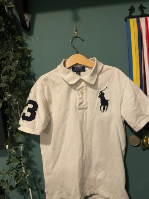 Vit piké från Polo Ralph Lauren S i barnstrl  - Vit pikétröja från Polo Ralph Lauren med klassisk krage, korta ärmar och stor mörkblå logga på bröstet. Snygg detalj med en stor siffra 3 på ena ärmen. Tillverkad i bomull och har en normal passform.Barnstorlek S
