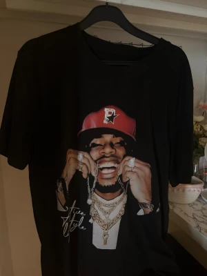 Svart t-shirt med tryck och kedjor - Cool svart t-shirt med stort tryck framtill av en person med röd keps, smycken och kedjor. T-shirten har korta ärmar och en avslappnad passform. Perfekt för dig som gillar streetstyle!