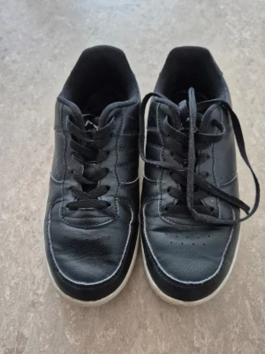 Sneakers - Snygga svarta sneakers från Nonation med klassisk design och vita sulor. Skorna har svarta snören, diskreta sömmar och ovandel i konstläder. Perfekta för en clean och stilren look till vardagsoutfiten.