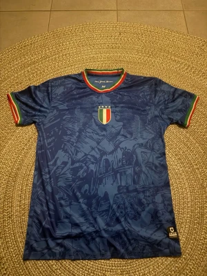 Blå Italien fotbollströja - Snygg blå fotbollströja från Givova med Italiens sköld på bröstet och detaljer i grönt, vitt och rött på krage och ärmslut. Tröjan har ett unikt mönster med italienska motiv och texten 'Italia' på ryggen. Perfekt för dig som älskar fotboll och vill sticka ut.