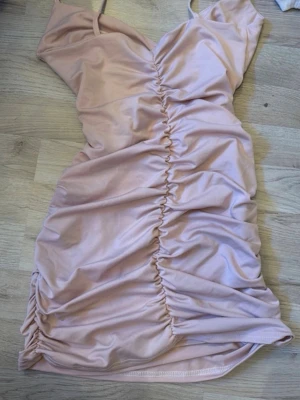 Ljusrosa omlottklänning med rynk - Trendig ljusrosa kortklänning med omlottdesign och rynkade detaljer längs hela framsidan. Klänningen har smala axelband och är tillverkad i ett mjukt, stretchigt material som ger en snygg passform. Perfekt för dig som gillar en feminin och modern stil.