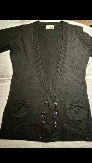 Svart kofta med rosetter från Koola Anna - Snygg svart kofta från Koola Anna of Sweden med djup V-ringning, dubbla rader knappar och två fickor med söta rosetter framtill. Ribbstickade muddar och mjukt stickat material gör den både stilren och bekväm. Perfekt för lager på lager och enkel att matcha.