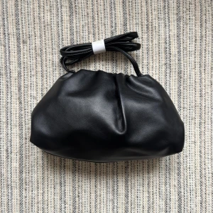 Svart clutchväska - Trendig svart clutchväska i mjuk veganskt läder med rynkad design och avtagbar axelrem. Väskan har en minimalistisk look och passar perfekt till dig som gillar stilrena accessoarer. Enkel att matcha med olika outfits.