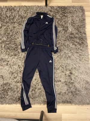 Marinblå Adidas träningsset - Säljer ett marinblått träningsset från Adidas med klassiska vita ränder längs ärmar och ben. Setet består av en zip-jacka och matchande byxor, båda med Adidas-logga på bröst och lår. Tillverkat i ett glansigt, sportigt material som är perfekt för träning eller chill.
