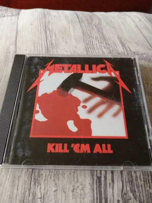 Metallica Kill 'Em All CD-album - Säljer Metallicas klassiska album 'Kill 'Em All' på CD. Omslaget är svart med röd text och grafik, och fodralet är i plast. Skivan innehåller låtar som 'Hit the Lights', 'The Four Horsemen' och 'Seek and Destroy'. Perfekt för dig som gillar hårdrock och samlar på musik.