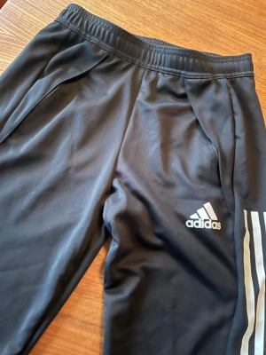 Svarta träningsbyxor från Adidas - Säljer ett par svarta träningsbyxor från Adidas med klassiska vita ränder längs benen och logga på låret. Byxorna har dragkedjor vid bensluten och elastisk midja. Perfekta för träning eller chill. Tillverkade i ett mjukt och stretchigt material.