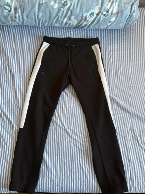 Svarta Lacoste mjukisbyxor med vita ränder - Säljer ett par svarta mjukisbyxor från Lacoste med vita ränder längs sidorna. Byxorna har dragsko i midjan, sidofickor och den klassiska Lacoste-loggan på vänster ben. Perfekta för chill dagar eller träning.