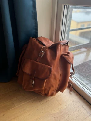 KERO ryggsäck i läder  - Snygg brun ryggsäck i äkta läder med klassisk design. Ryggsäcken har ett stort huvudfack, en ficka framtill med spänne och justerbara axelband. Perfekt för dig som vill ha en stilren och praktisk väska till skolan eller stan. Ny pris 3390kr. Allt i dm 