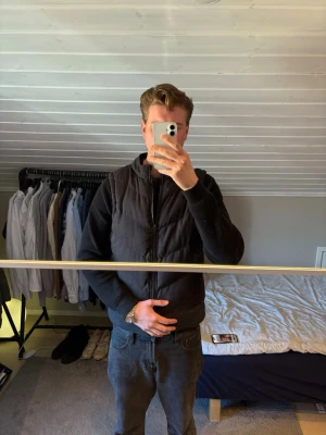 Svart dunväst med dragkedja - Snygg svart dunväst med dragkedja framtill och hög krage. Västen har en stilren och enkel design som passar till det mesta. Perfekt att slänga över en hoodie eller tröja för extra värme. Materialet är quiltat och ger en modern look.