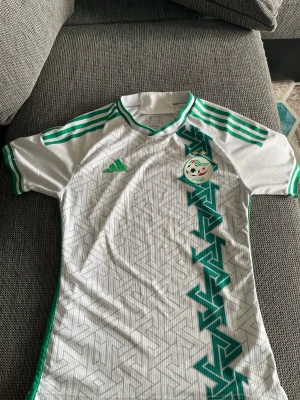 Vit och grön Algeriet fotbollströja Adidas - Säljer en snygg vit och grön fotbollströja från Adidas med Algeriets landslagsmärke på bröstet. Tröjan har geometriskt mönster, gröna detaljer och tre klassiska Adidas-ränder på axlarna. Perfekt för dig som gillar fotboll och vill representera Algeriet.