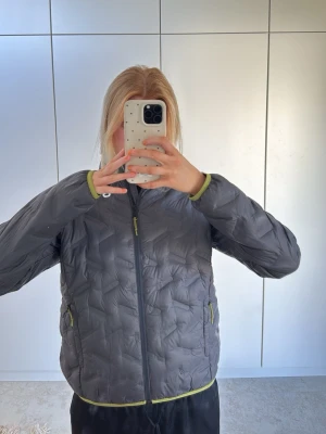Mörkgrå pufferjacka från 8848 Altitude - Snygg mörkgrå pufferjacka från 8848 Altitude med limegröna detaljer på dragkedjor och kanter. Jackan har hög krage, två sidofickor med dragkedja och quiltad design. Perfekt för kalla dagar och riktigt skön att bära.