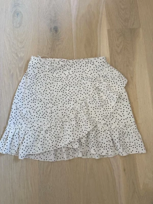 Vit prickig omlottkjol med volang - Supersöt vit kort volangkjol med svarta prickar och omlottdesign. 100% polyester. Storlek s