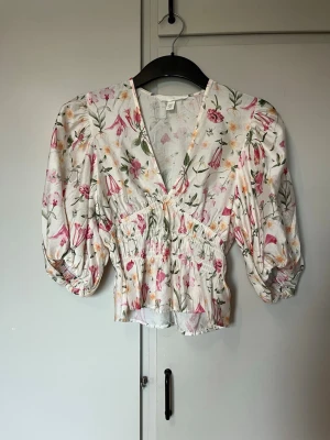 Blommig v-ringad blus med puffärm - Söt blus med v-ringning och puffiga trekvartsärmar. Vit botten med blommigt mönster i rosa, grönt och gult. Smockad midja. 