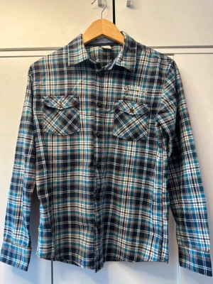 Blå rutig skjorta med fickor - Snygg blå och vit rutig skjorta med långa ärmar och två bröstfickor med lock. Skjortan har klassisk krage och knäppning framtill. Perfekt för en avslappnad och cool stil.