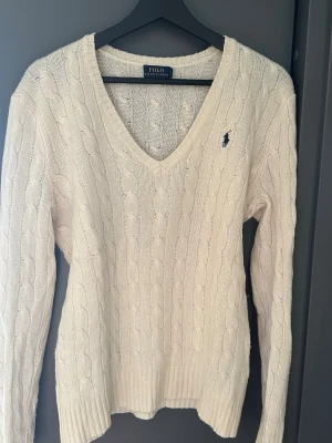 Vit kabelstickad tröja Polo Ralph Lauren - Kabelstickad vit tröja från Polo Ralph Lauren med djup V-ringning och klassisk marinblå logga broderad på bröstet. Tröjan har långa ärmar och ribbade muddar. Perfekt för dig som gillar tidlös och clean stil. 90% merinoull och 10% Kashmir 
