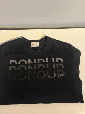 Svart sweatshirt från Dondup - Cool svart sweatshirt från Dondup med stor, glansig logga tryckt framtill i flera lager. Bra skick men nedre ärmen är lite smutsig men kan gå bort i tvätten. Tröjan har rund halsringning och långa ärmar med ribbade muddar. Tillverkad i mjukt bomullsmaterial som känns skönt mot huden. Perfekt för en avslappnad och trendig look.