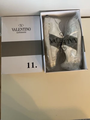 Vita sneakers från Valentino Garavani - Snygga vita sneakers från Valentino Garavani med klassisk låg siluett och diskret grå detalj på sidan. Skorna har en stilren design och är tillverkade i läder med vita snören. Perfekta för dig som gillar exklusiva märken och minimalistisk stil.
