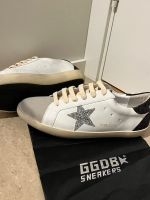 GGDB Superstar vita sneakers - Snygga vita GGDB Superstar sneakers med silverglittrig stjärna på sidan, beigea skosnören och grå mockadetalj vid tån. Baksidan har svart glitter och sulan är beige. Tillverkade i läder och mocka, perfekta för dig som gillar en unik och trendig look.ä Golden Goose goldengoose skor sneakers märkeskläder yk2 grisch sommar billigt sommarkläder sommarskor