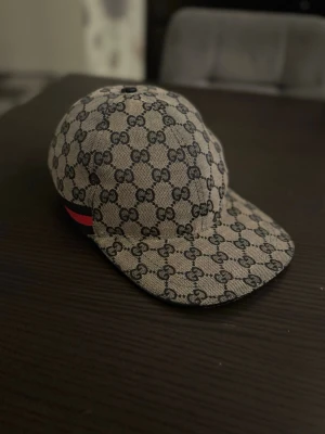  Gucci Keps   - Säljer en snygg keps från Gucci med klassiskt GG-monogram i beige och mörkblått. Kepsen har en böjd skärm och justerbart band bak. Materialet är canvas och på sidan finns en röd och marinblå rand som ger extra lyxig känsla. KAN GÅ NER I PRIS VID SNABBAFFÄR
