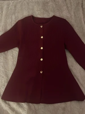Vinröd ribbad kofta med guldknappar - Snygg vinröd kofta med ribbad struktur och rund halsringning. Koftan har långa ärmar och stängs med fem guldfärgade knappar framtill. Passar perfekt till lager på lager och ger en elegant touch till din outfit.