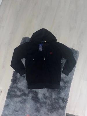 Svart hoodie från Polo Ralph Lauren - Svart hoodie med dragkedja från Polo Ralph Lauren. Klassisk design med huva och fickor framtill. Liten röd logga broderad på bröstet. Tillverkad i mjukt bomullsmaterial som är skönt att bära. Perfekt för en avslappnad och stilren look.