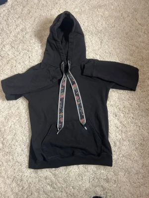 Svart hoodie med blommiga snören - Cool svart hoodie med stor huva och unika breda snören med blommönster. Perfekt för dig som vill ha en enkel men ändå personlig stil. Materialet känns mjukt och bekvämt, troligen bomull eller bomullsblandning.