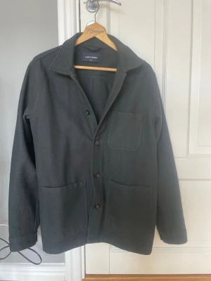 Overshirt från A Day's March - Stilren mörk overshirt från A Day's March med klassisk krage, tre utanpåliggande fickor och knäppning framtill. Tillverkad i ett mjukt, tjockare tyg som ger en avslappnad men ändå snygg look. 