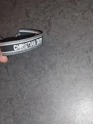Christian Dior J'Adior tygarmband - Snyggt svart och vitt tygarmband från Christian Dior med texten 'J'Adior'. Armbandet har flätade band och tofsar i ändarna, samt detaljerade kanter med små vita prickar. Perfekt accessoar för att lyfta din outfit.