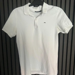 Vit pikétröja från J.Lindeberg - Klassisk vit pikétröja från J.Lindeberg med liten logga på bröstet. Tröjan har krage, korta ärmar och knappar vid halsen. Perfekt till en clean och stilren look.