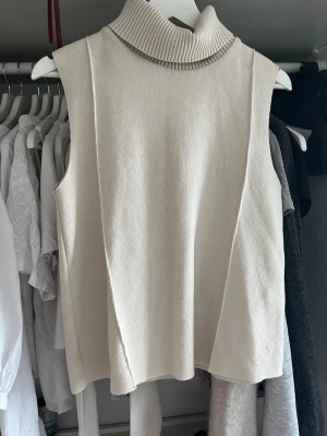 Ärmlös beige polotröja - Säljer en stilren ärmlös polotröja i beige stickat material. Tröjan har hög, ribbad krage och en rak, avslappnad passform. Perfekt att styla över en skjorta eller bära som den är för en clean look.