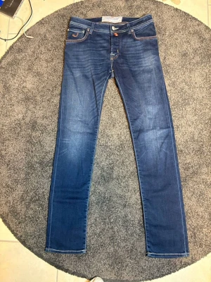 Blå jeans från Jacob Cohën - Snygga blå jeans från Jacob Cohën med klassisk femficksmodell och kontrastsömmar. Jeansen har en orange patch med ett stort J på bakfickan och tydliga vita sömmar. Perfekta för en trendig och avslappnad stil.