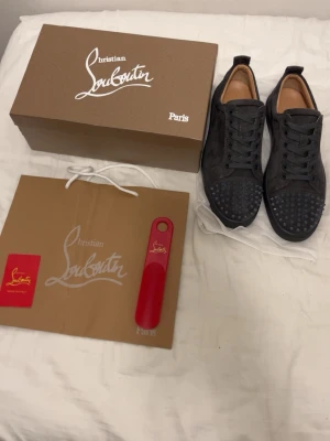 Gråa Louboutin sneakers med nitar - Säljer ett par gråa sneakers från Christian Louboutin i mocka med matchande grå snörning och coola nitar över tån. Skorna har en låg siluett och den klassiska exklusiva Louboutin-looken. Perfekta för dig som vill sticka ut med stil.