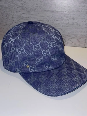 Blå GG-mönstrad keps från Gucci - Snygg blå keps från Gucci i denim-look med det klassiska GG-mönstret över hela kepsen. Böjd skärm och justerbar baksida. Perfekt för dig som vill ha en trendig accessoar med lyxig känsla och tydlig logga.