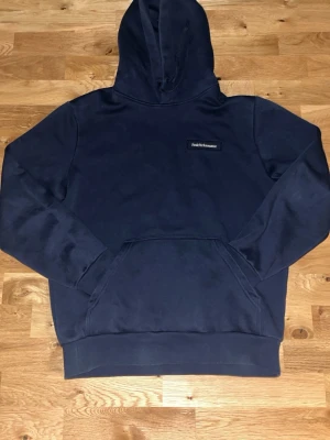 Mörkblå hoodie från Peak Performance - Snygg mörkblå hoodie från Peak Performance med broderad logga på bröstet. Klassisk modell med huva och stor magficka framtill. Perfekt för en avslappnad och stilren look. Mjuk bomullsblandning som känns skön mot huden.