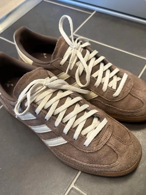 Bruna Adidas Gazelle handball spezial  sneakers 42.5 - Säljer ett par bruna Adidas Gazelle sneakers i mocka med klassiska vita ränder på sidorna och vita skosnören. Skorna har en låg profil och gummisula som ger en retrovibe. Perfekta för dig som gillar stilrena och tidlösa sneakers.