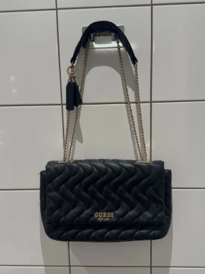 Svart handväska från Guess - Snygg svart handväska från Guess med quiltad design och guldfärgade kedjor som axelrem. Väskan har Guess-logga i guld framtill och ett dekorativt hänge. Perfekt accessoar för att lyfta din outfit.