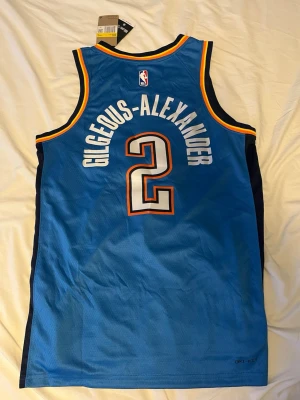 SGA Oklahoma City Thunder basketlinne - Säljer ett blått basketlinne från Nike med Oklahoma City Thunder-tryck och nummer 2, GILGEOUS-ALEXANDER på ryggen. Linnet har vita och orange detaljer, är ärmlöst och tillverkat i ett lätt, ventilerande material perfekt för basket eller träning.