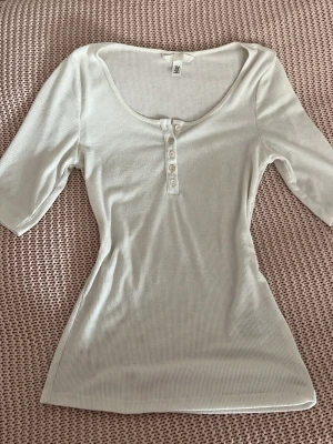 Vit ribbad topp med knappar - Säljer en vit ribbad topp från H&M med rundad halsringning och knappdetaljer framtill. Toppen har halvlånga ärmar och är figurnära i modellen. 