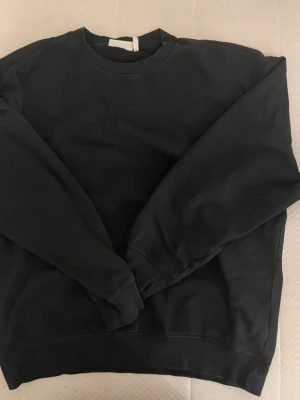 Svart basic sweatshirt - En enkel svart sweatshirt med rund hals och långa ärmar. Tröjan har en avslappnad passform och ribbade muddar vid ärmslut och nederkant. Perfekt för en casual look och lätt att matcha med andra plagg.