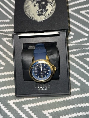 Blå och guld klocka Versus Versace - Snygg klocka från Versus Versace med blå silikonarmband och blå urtavla. Boetten är i guldtonad metall och har flera funktioner på urtavlan. Klockan levereras i originalbox med Versus-logga. Perfekt accessoar för dig som vill sticka ut. OBS! Klockan har stannat