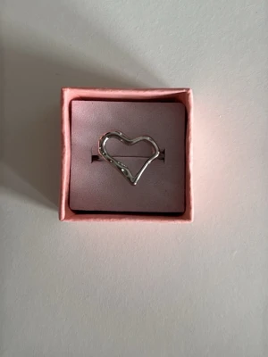 Silverfärgad hjärtring i metall - Säljer en stilren ring i silverfärgad metall med ett öppet hjärta som motiv. Ringen har en modern, enkel design och levereras i en söt rosa ask. 