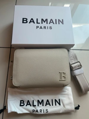 Beige axelväska från Balmain - Stilren beige axelväska från Balmain i strukturerat läder med präglad B-logga framtill. Väskan har en bred justerbar axelrem och levereras med original dustbag och box. Perfekt för dig som vill ha en exklusiv och trendig accessoar.